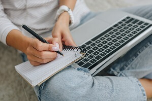 Ausgeschnittene Ansicht einer Frau, die in Notizbuch schreibt und Laptop hält